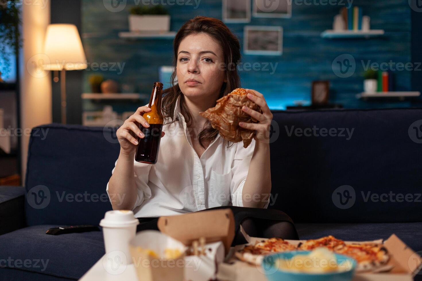 Woman after office work eating tasty takeaway hamburger holding beer bottle on couch watching news on television. Serious person having takeout burger and fast food delivery menu. photo