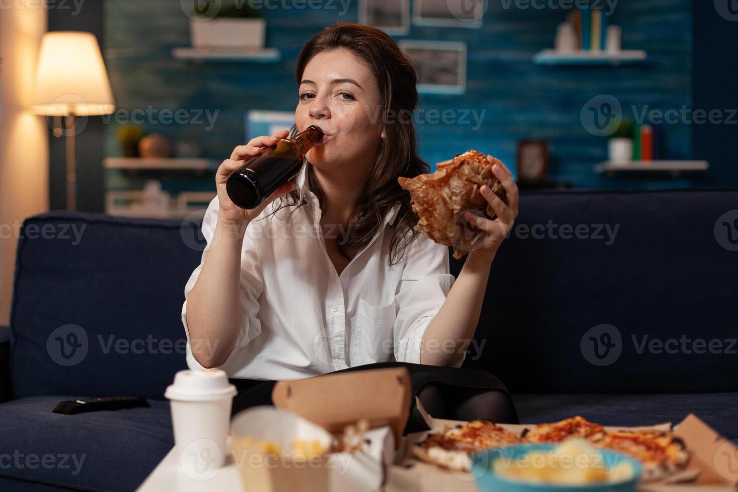 Woman in white shirt drinking beer from bottle while holding a tasty takeaway burger looking at television movie. Person sitting on couch eating delivery hamburger in front of table with fast food. photo