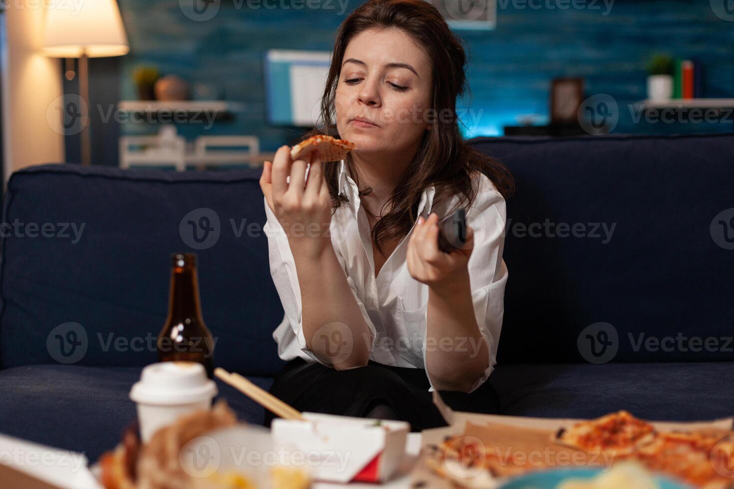 Woman looking at a slice of home delivered pizza while holding remote switching channels on television in front of coffee table with takeaway menu. Person having fast food browsing tv stations. photo