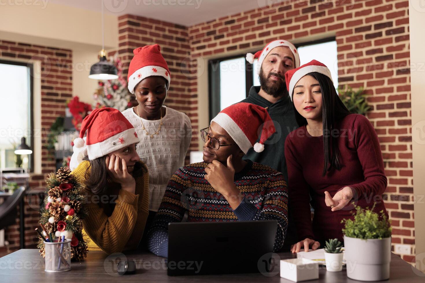 African american, caucasian, and asian workers wearing santa hats discussing task on christmas eve. Startup company colleagues managing project on laptop together in festive decorated office photo