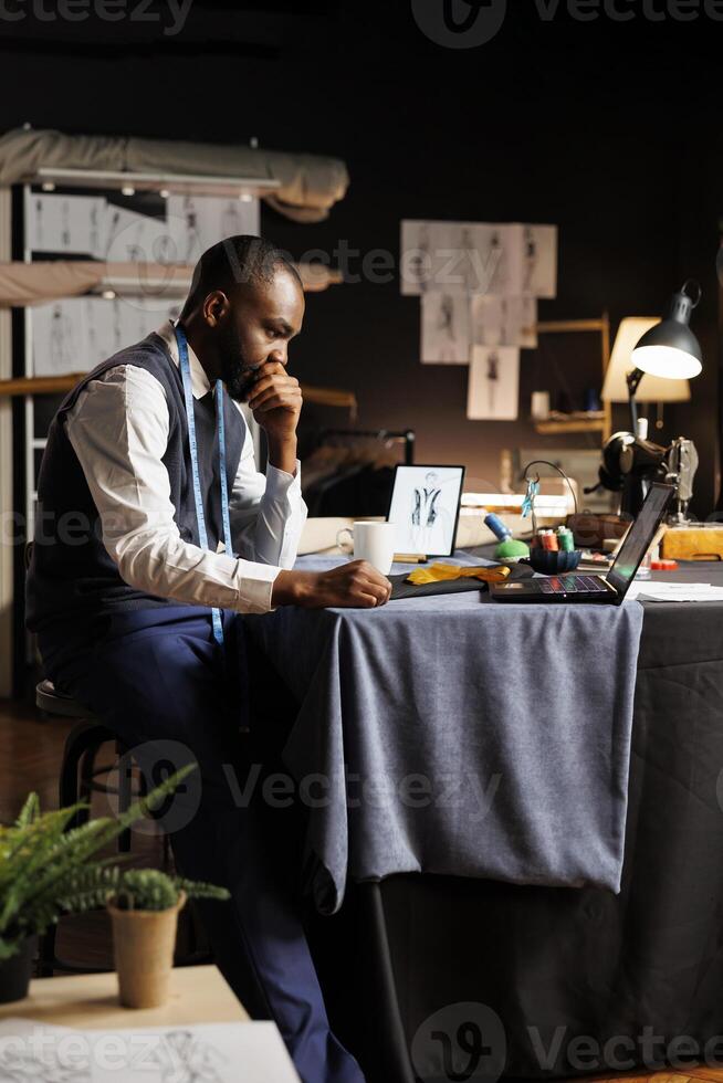 Couturer looking online for inspiration, struggling to find ideas for upcoming fashion design runway show collection. Suitmaker working on bespoken sartorial garments in atelier shop workspace photo