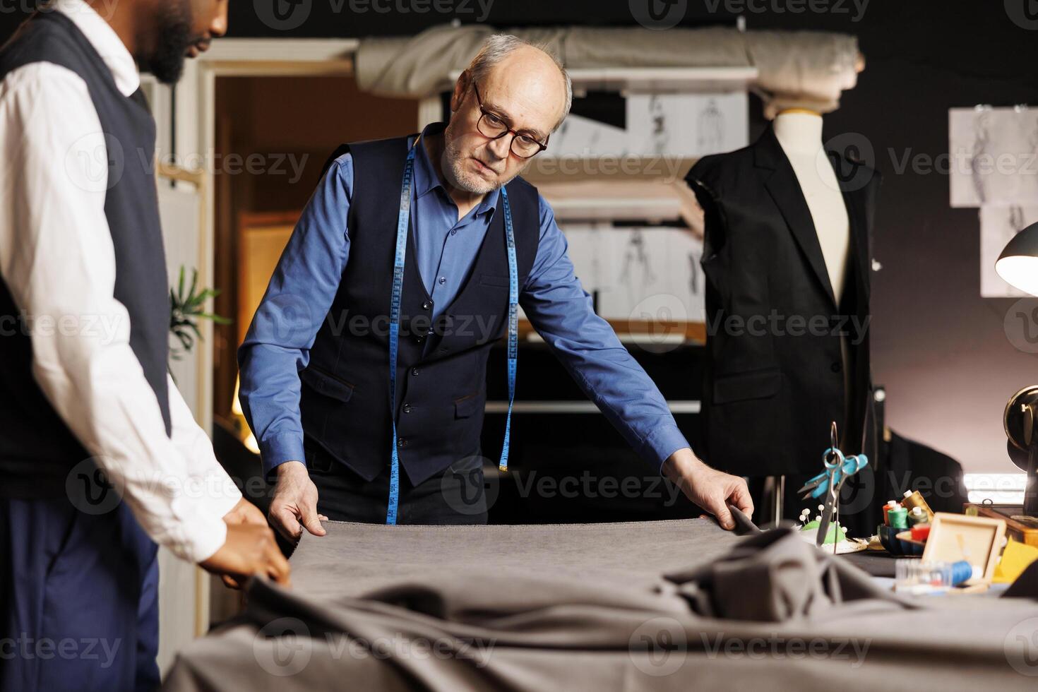 Experienced elderly master suitmaker preparing new fabric sheet for upcoming sartorial collection manufacturing. Senior tailor and apprentice ready to measure, cut, sew and stich textile material photo