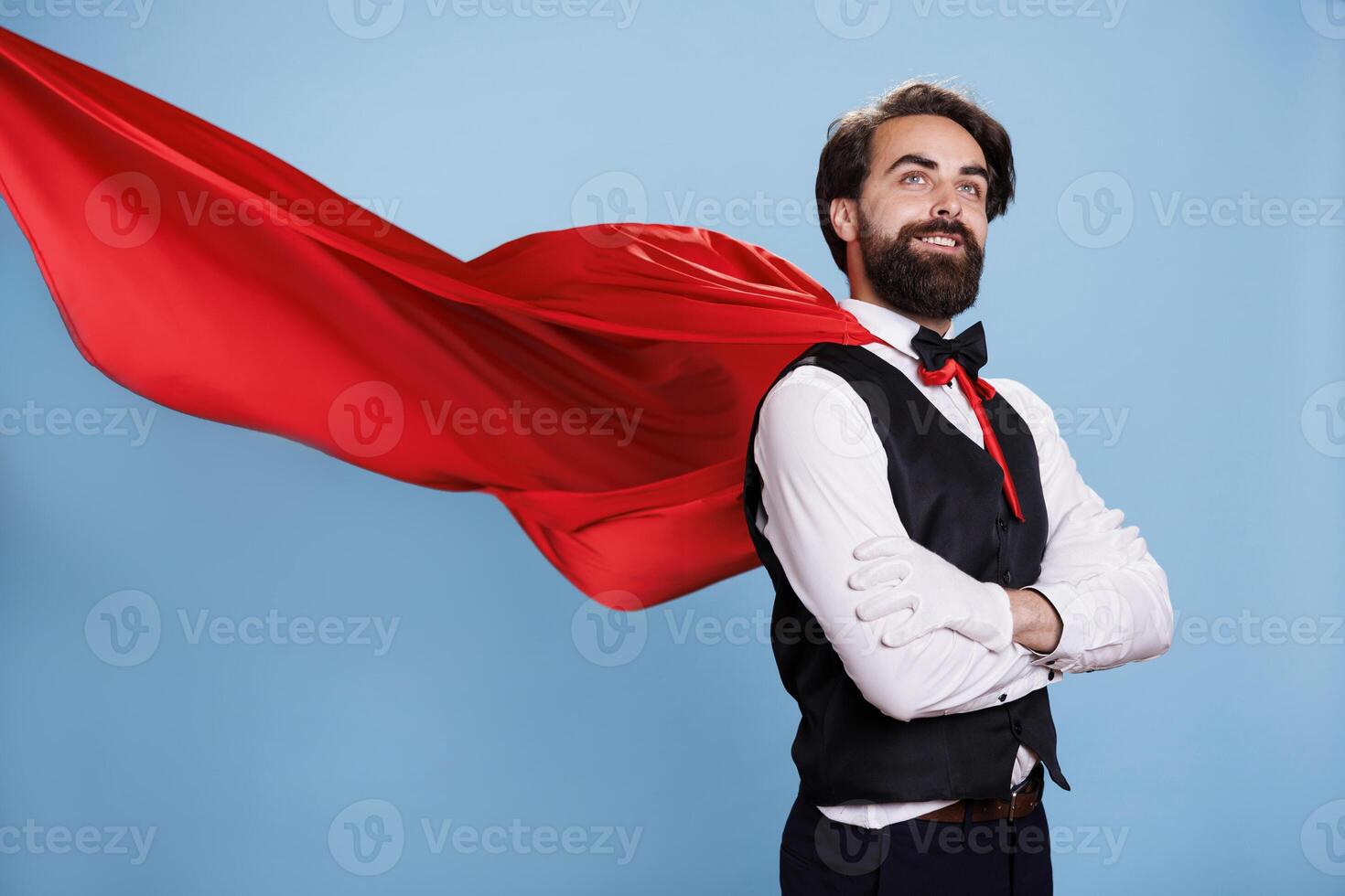 Determined person standing in studio, red cape waving around against blue background. Young adult in suit and tie coming to rescue citizens, pretending to be a superhero in fantasy cartoon. photo