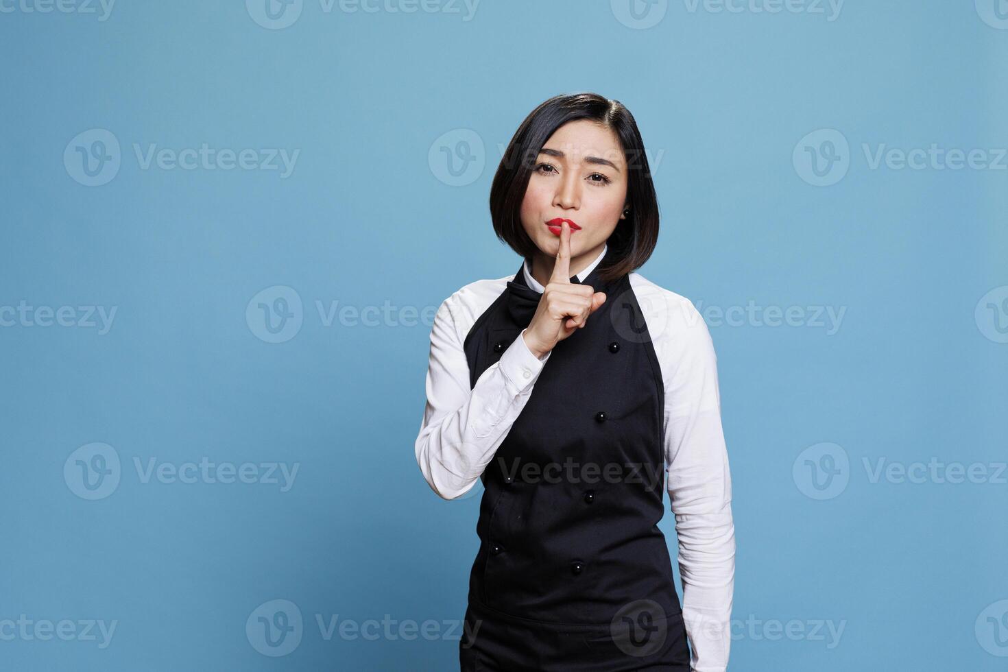 Asian waitress dressed in restaurant uniform showing silent gesture and looking at camera with serious face expression. Woman receptionist making tsss sign while holding finger on lips portrait photo