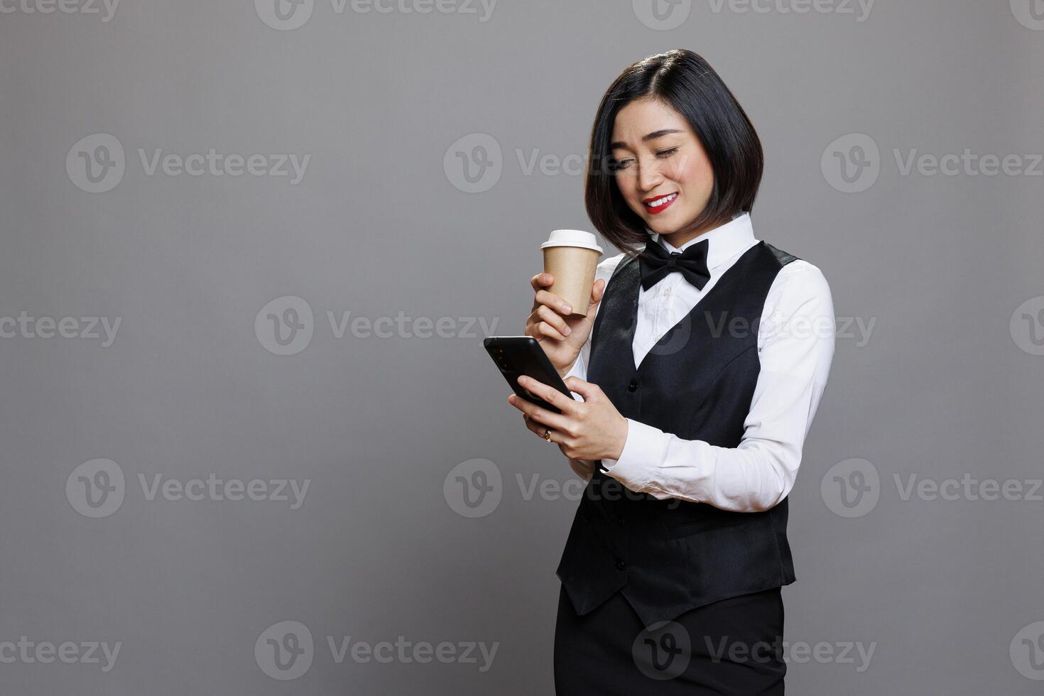 Carefree asian woman in waitress uniform drinking coffee and using social media app on smartphone. Catering service smiling worker holding tea to go paper cup and communicating online on mobile phone photo