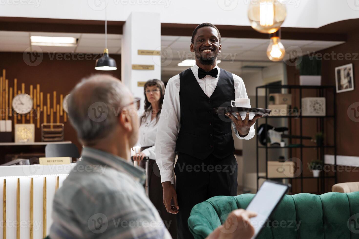 In luxury hotel lobby, african american waiter serves coffee to elderly gentleman visitor using digital tablet. In lounge area, retired senior guy holds device and accepts a cup of tea from employee. photo