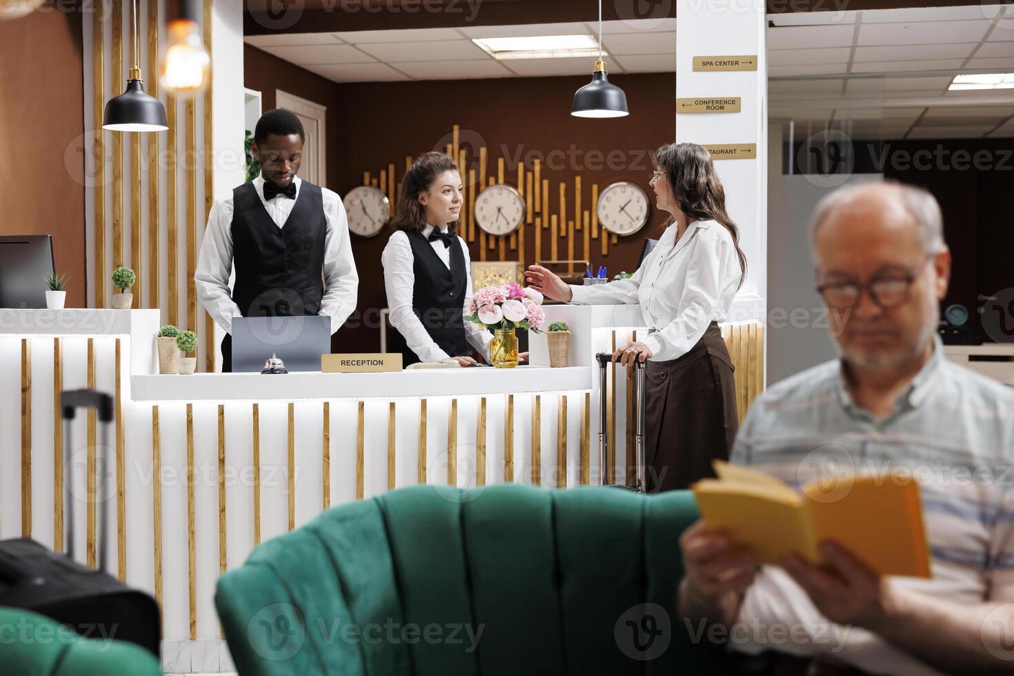 With assistance from receptionist in hotel lobby, retired senior woman with suitcase checks in at front desk. While female visitor waits at counter, an elderly man reading in the cozy lounge. photo