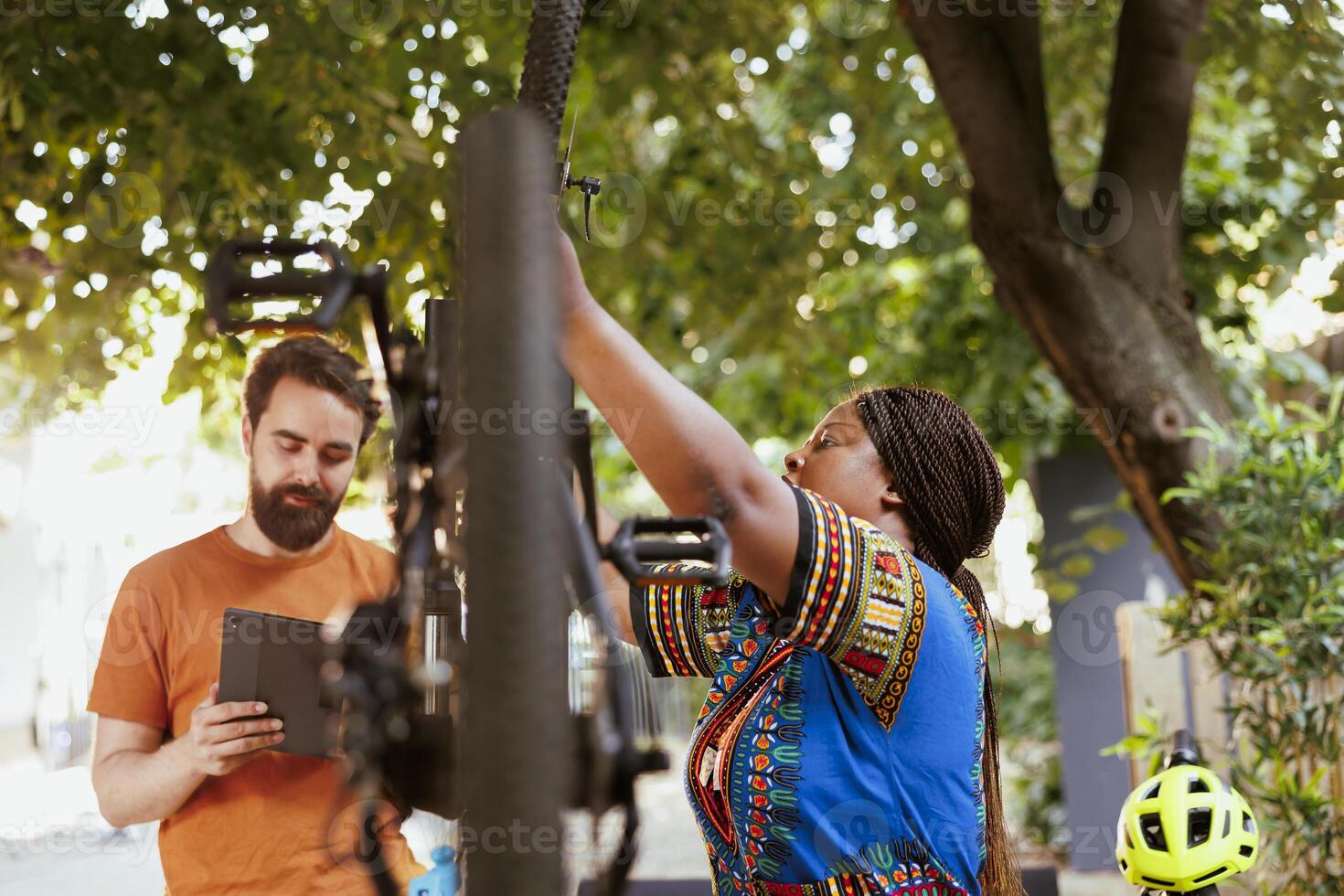 Caucasian man standing with digital device while african american woman repairs damaged bike. Multiethnic couple using phone tablet for researching of bicycle adjustments for summer maintenance at home yard. photo