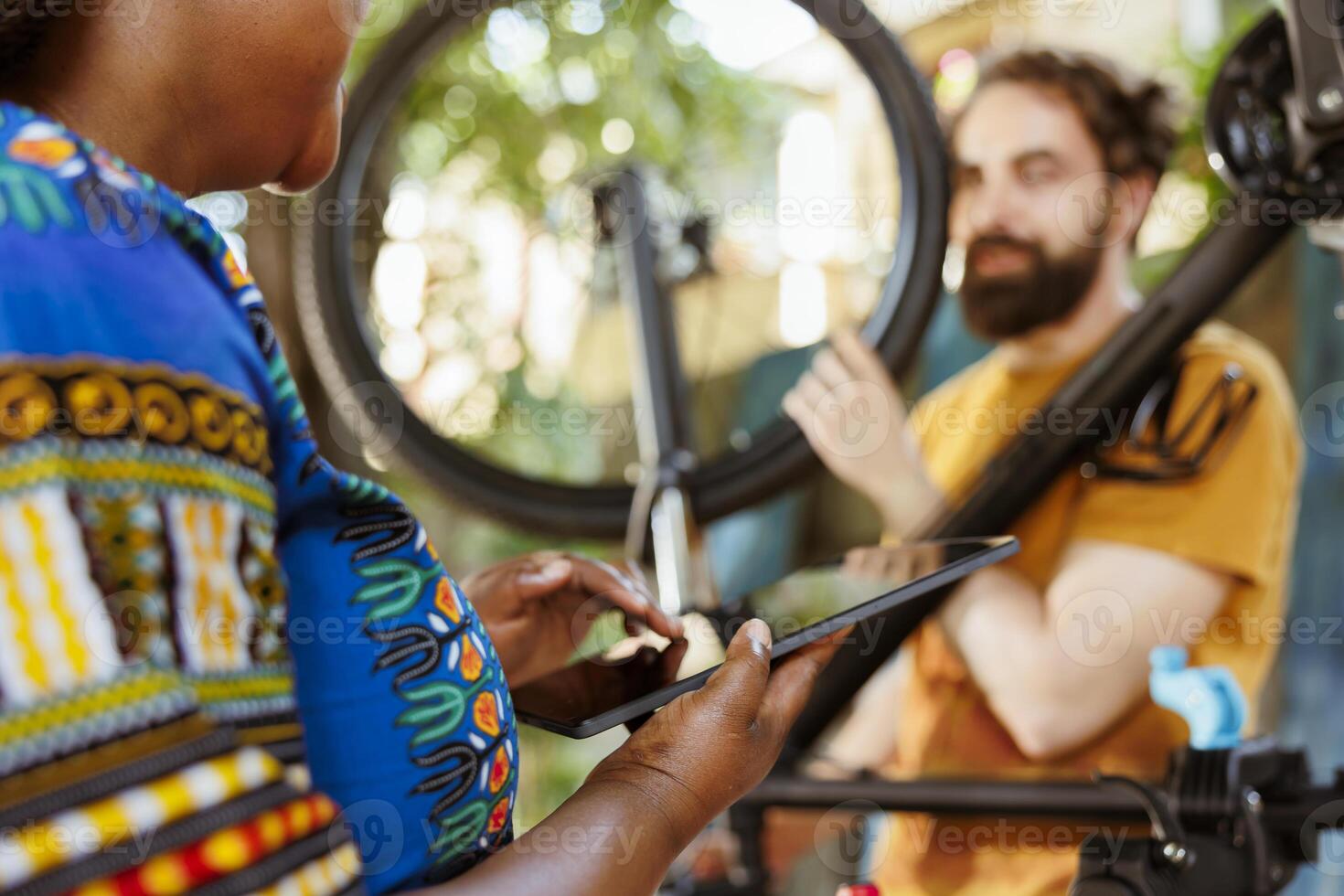 Young african american female holding phone tablet while man in background inspects bicycle wheel outside. Black woman using smart digital device to look for solutions of damaged bike tire. photo