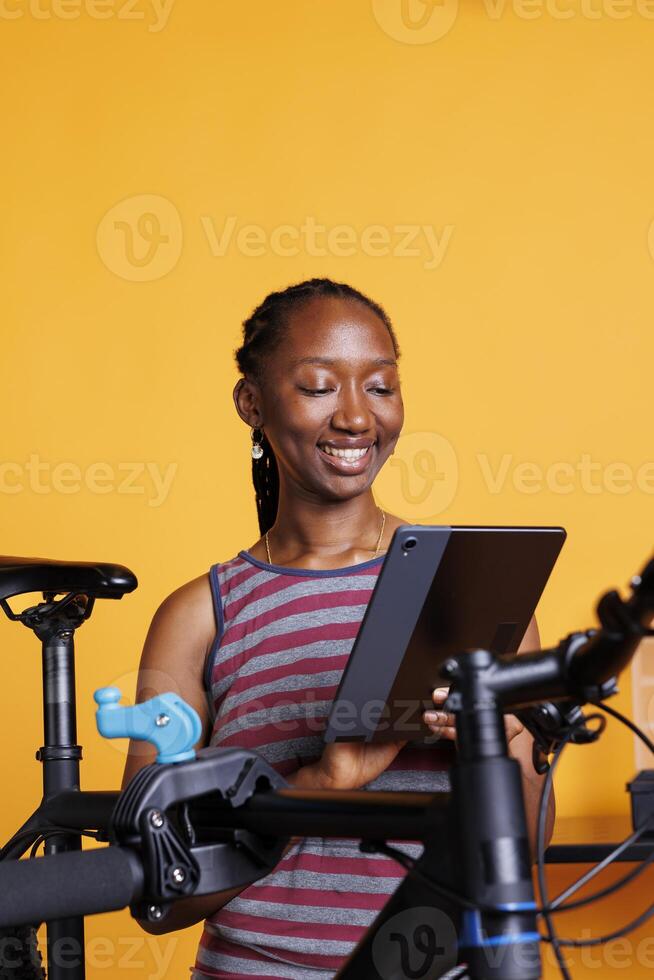 Active sporty woman using smart digital device to research bike repair solutions. Smiling african american female holding phone tablet and checking for damages on bicycle, against isolated background. photo