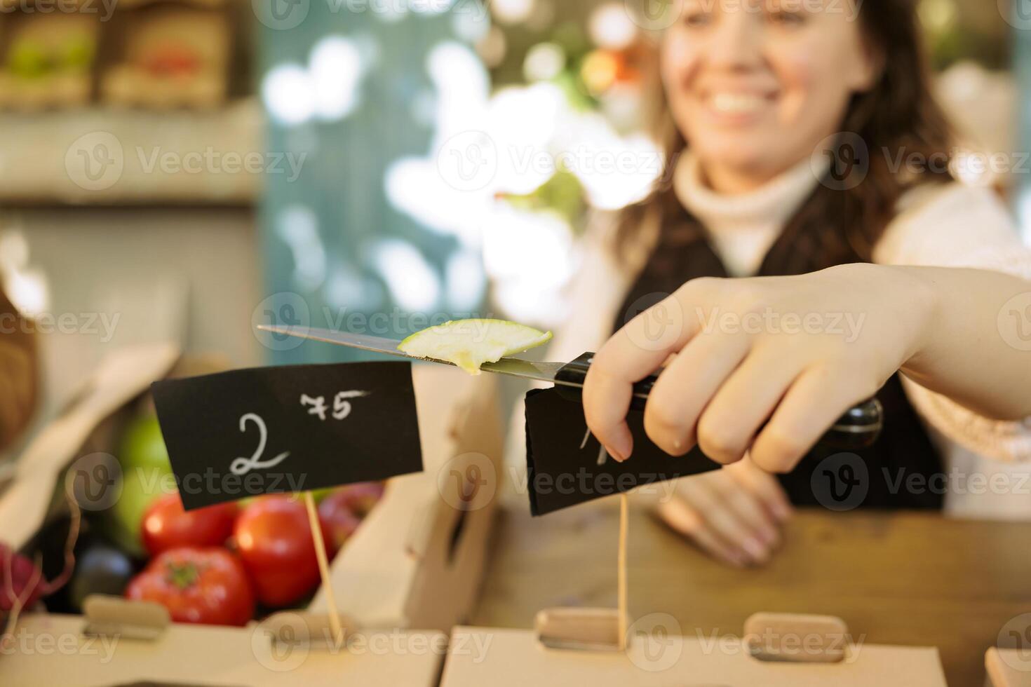 Close up of young farmer cutting fruits or veggies organic produce, healthy food at local farmers market. Selective focus on female vendor holding knife having a piece of fresh bio apple. photo