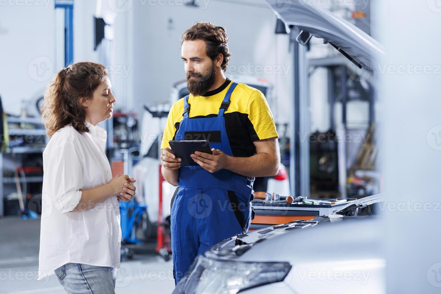Repairman at auto repair shop conducts annual vehicle checkup, informing client about needed engine replacement. Proficient garage worker talks with customer after finishing car inspection photo