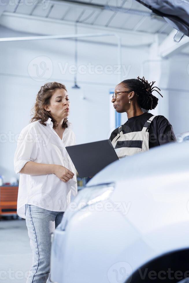African american mechanic in car service uses laptop to calculate invoice after fixing broken automobile engine. Garage worker uses device to inform woman of final costs after mending vehicle photo