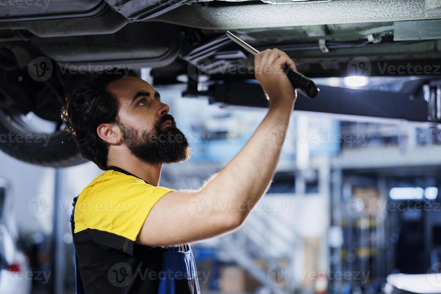 Qualified engineer uses lamp light to asses situation before changing old car brake fluid with new one in repair shop. Meticulous worker in garage doing checkup on vehicle suspended on lift photo