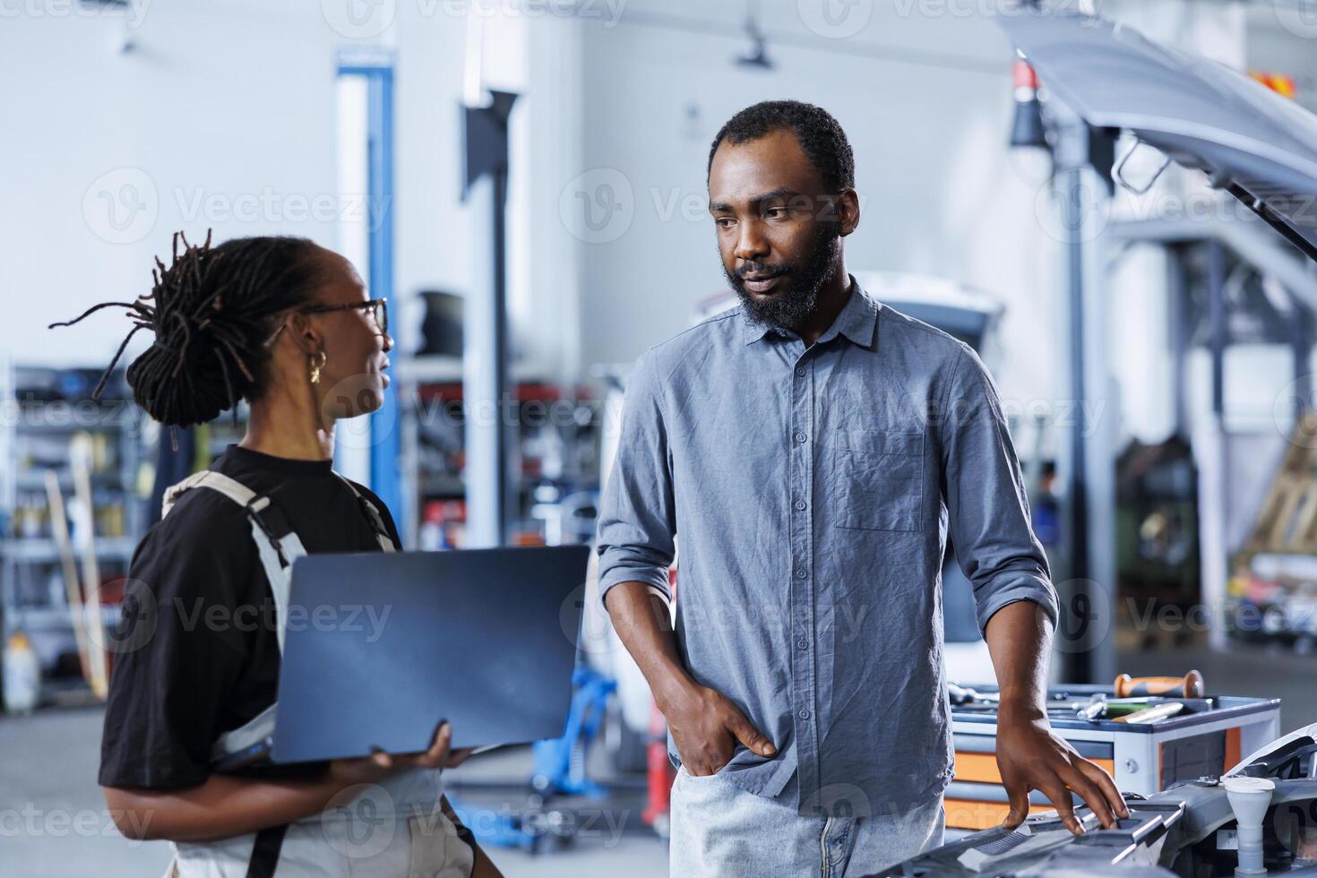 BIPOC woman next to client in car service orders new engine for damaged vehicle using laptop. Garage employee looking for components on internet to replace old ones in faulty automobile photo