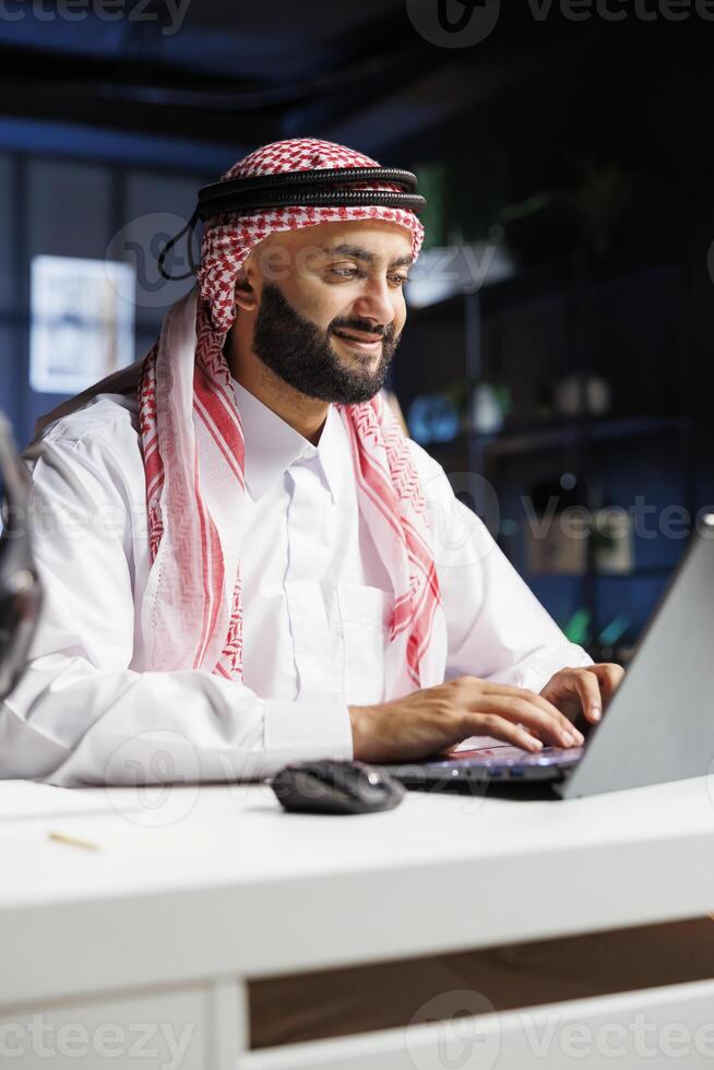While seated at his home office desk, a Middle Eastern male entrepreneur works effectively on his laptop. Portrait of Arab man checking e-mail, perusing the internet, and thoroughly examining notes. photo