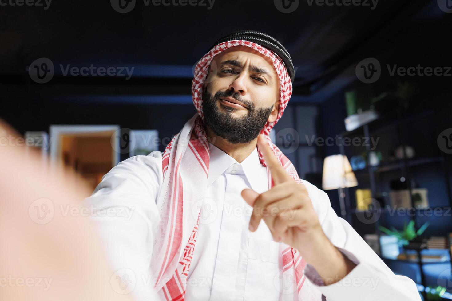 POV of a jovial Muslim man participating in a conference. Middle Eastern guy wearing traditional clothing is vlogging online while holding a smart gadget and pointing at the camera. photo