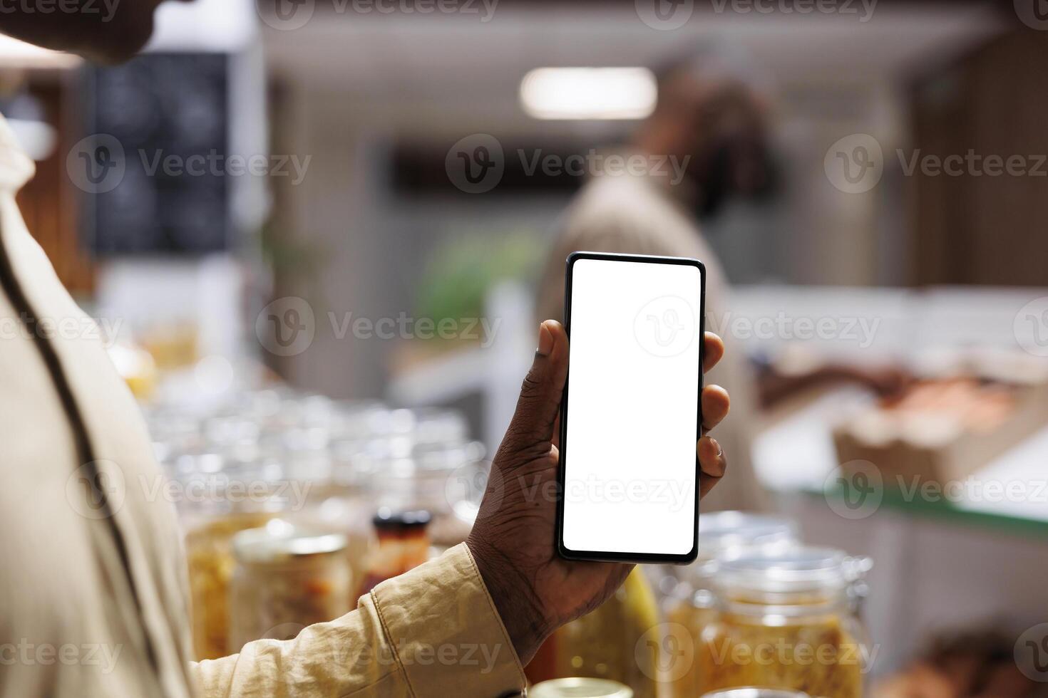 Close-up shot of African American male customer vertically holding a mobile device with an isolated chromakey mockup template. The store sells fresh, eco friendly items on wooden shelves. photo