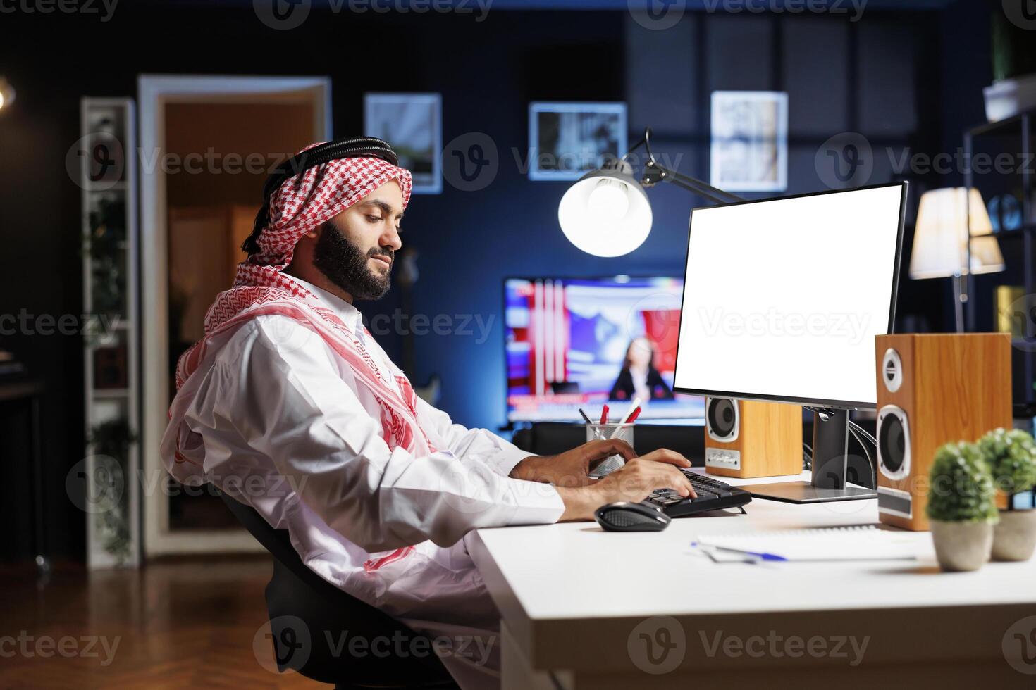 Side-view of Arab man sitting at the table with his pc monitor displaying an isolated chromakey template. Muslim guy working on a computer desktop with a blank mockup white screen. photo