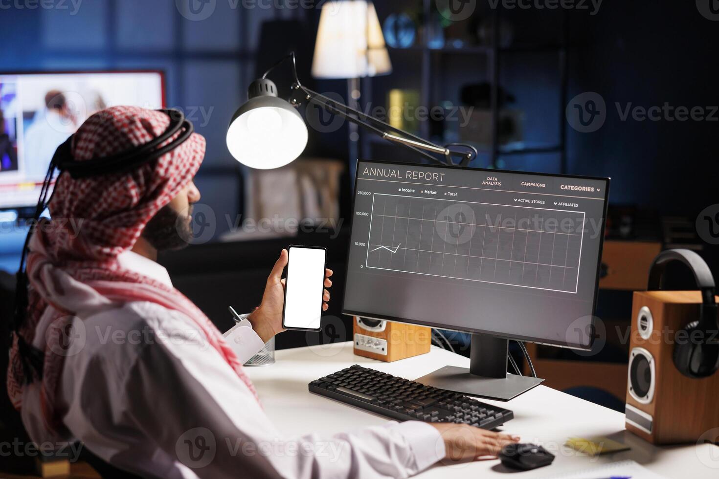 Male person in traditional attire sits at desk presenting a promo on mobile device with isolated copyspace template. The individual focus on phone reflects the integration of technology and culture. photo