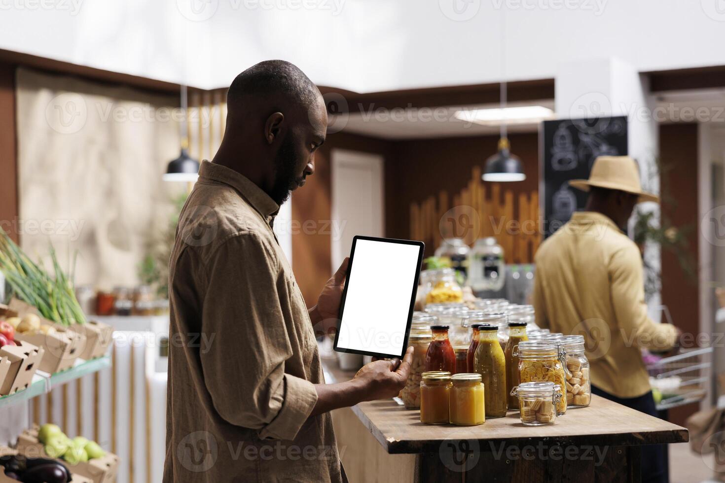 African American man checking the eco friendly products, jars, and groceries on shelves while holding a tablet with a chromakey template. Blank screens for mockups. Sustainable local supermarket. photo