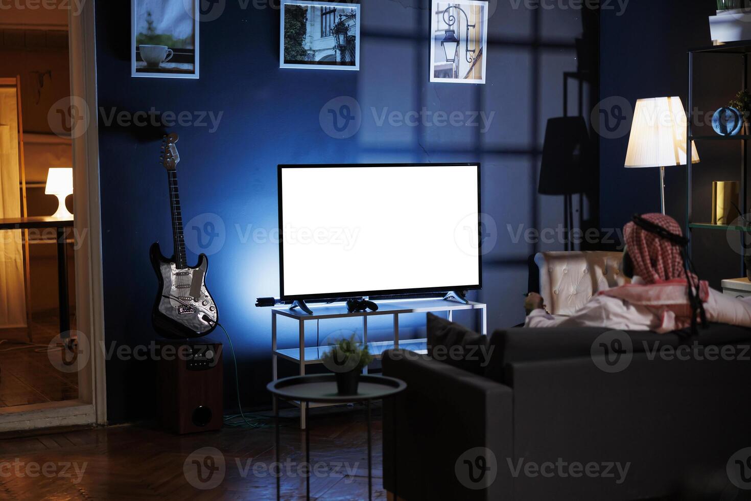 Back-view shot of Muslim guy in traditional attire seated on sofa in front of television displaying isolated white screen. Image showing man looking at tv screen with blank copyspace mockup template. photo