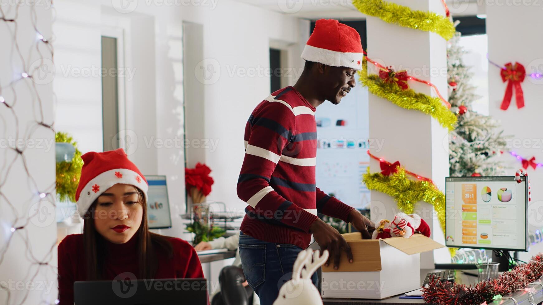 Exicted african american employee clearing desk before leaving for vacation, dancing around festive decorated office. Man wearing Santa hat taking days off to go on xmas holiday, feeling merry photo