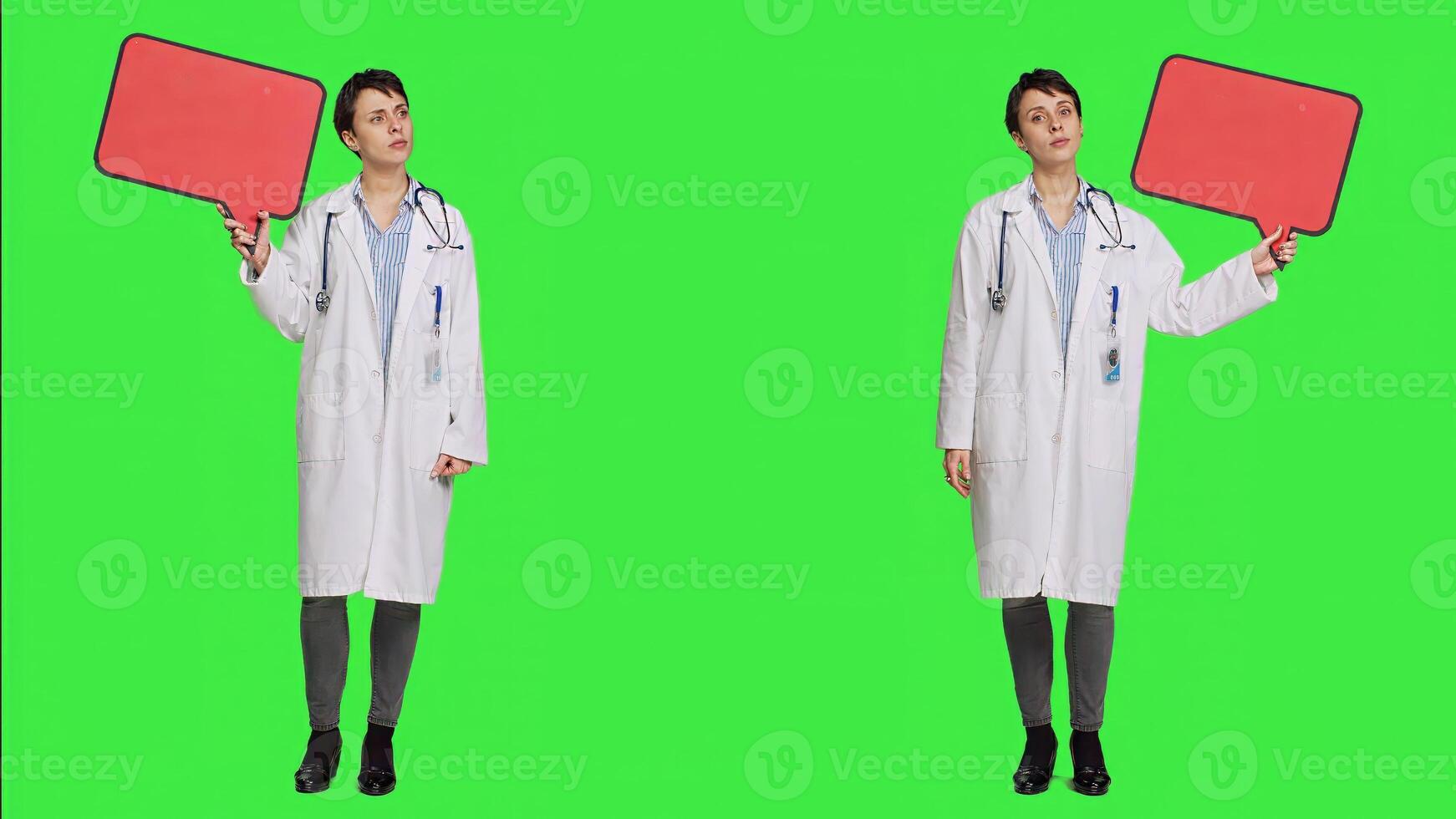 Woman doctor holding a red empty speech bubble sign in studio, showing an isolated blank cardboard icon against greenscreen backdrop. Physician creating announcement with banner. Camera A. photo