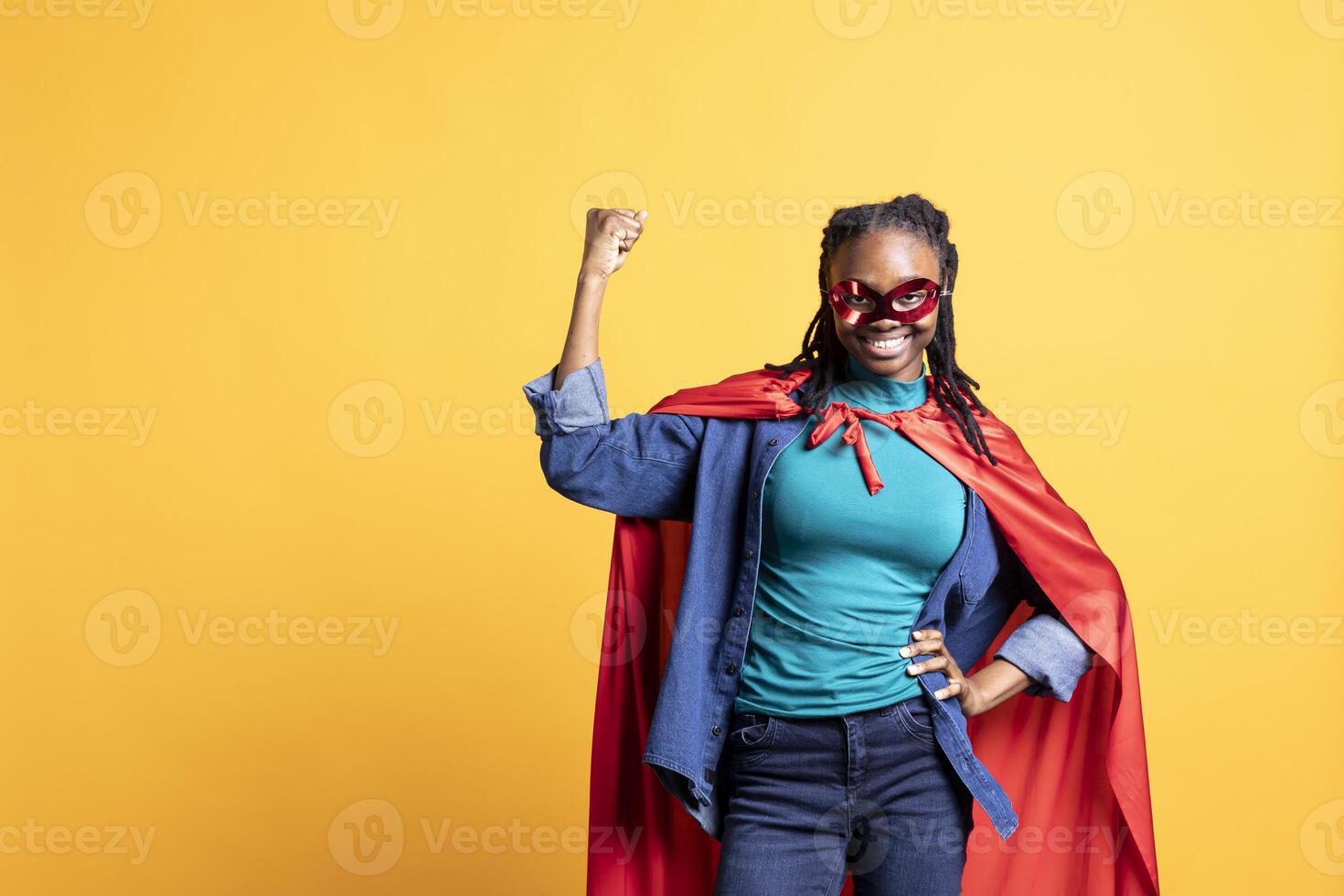 African american girl portraying superhero with red cape, isolated over studio background, flexing muscles. Smiling teenager wearing cloak posing as hero in costume, having fun photo