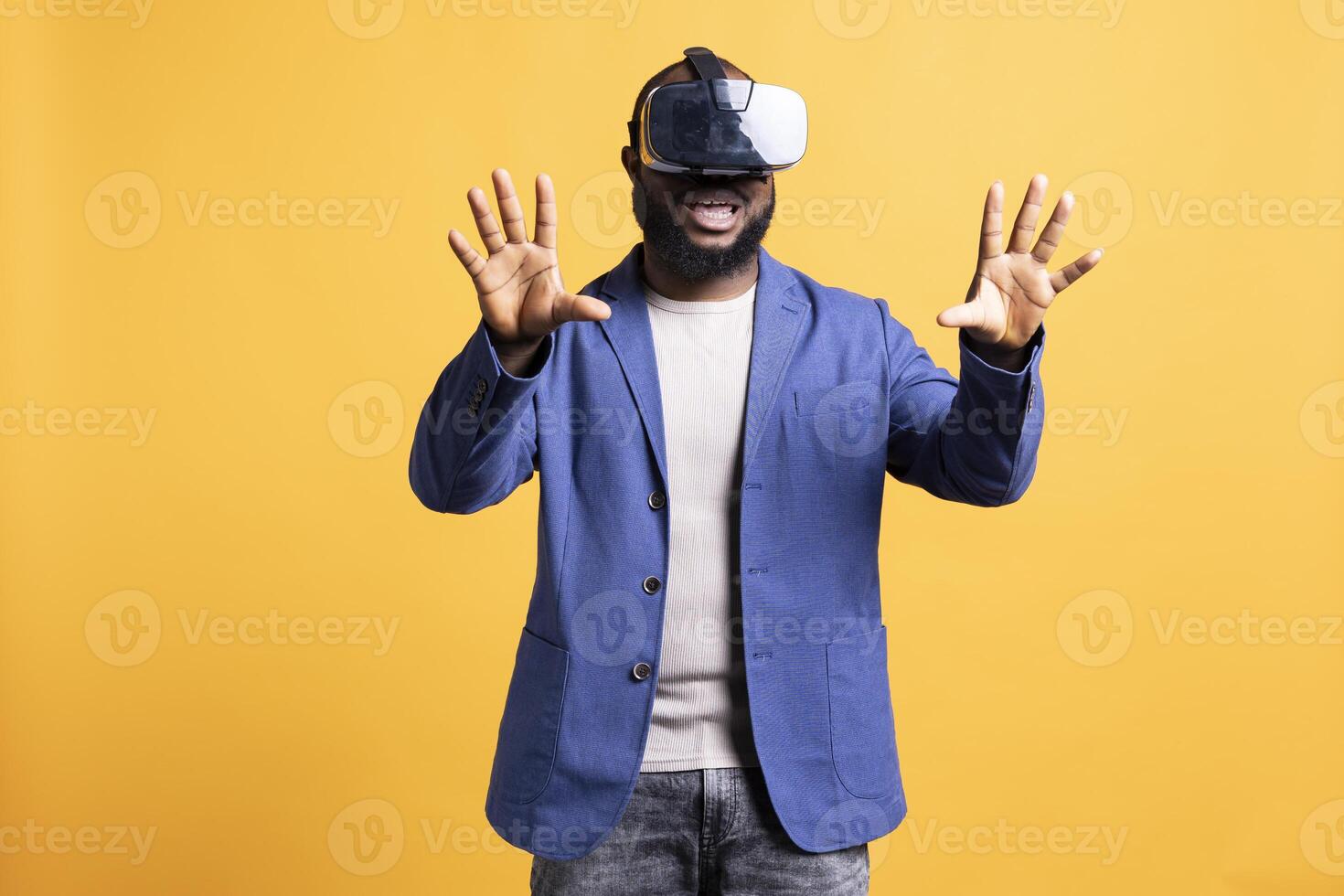 Joyous african american man wearing virtual reality headset, interacting with interface. Delighted person having fun using high tech futuristic modern VR gadget, isolated over yellow studio background photo