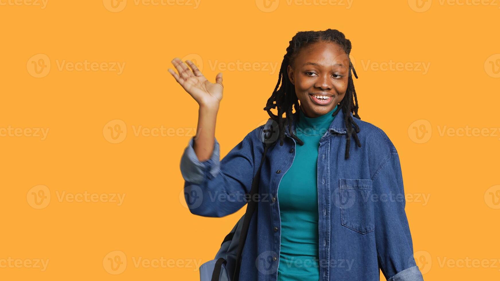 Portrait of happy woman waving hand, saluting, gesturing, isolated over yellow studio background. Cheerful BIPOC teenager with school rucksack doing greeting hand gesture, camera A photo