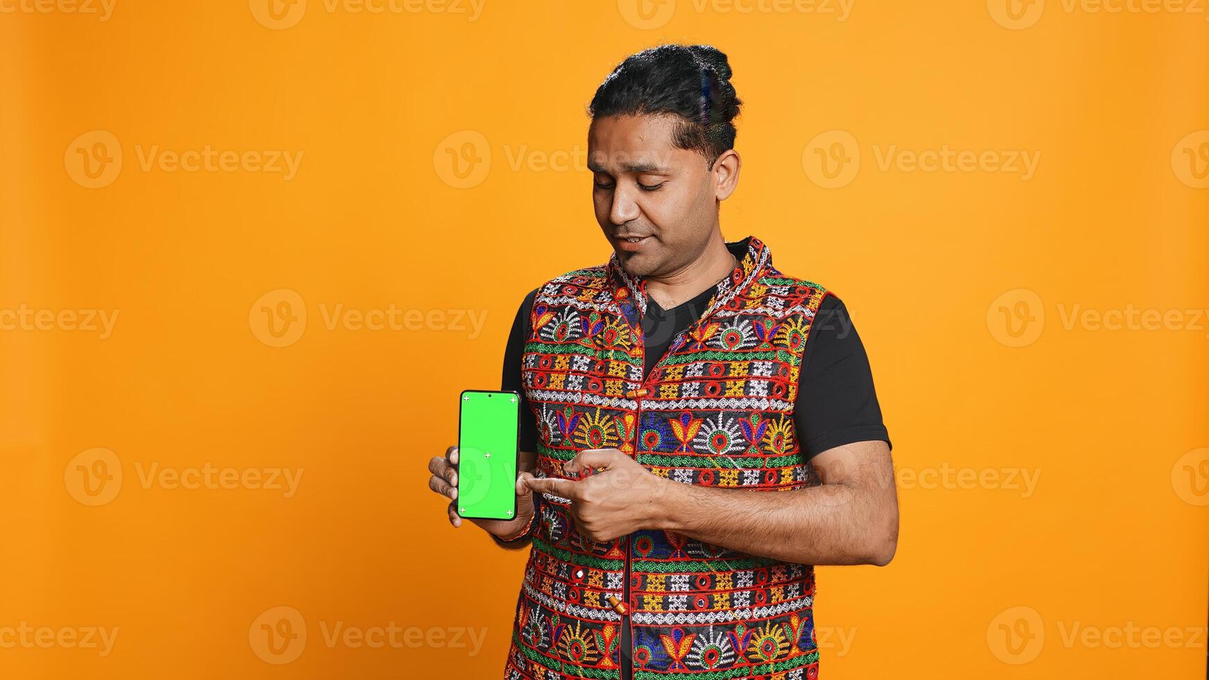 Influencer presenting green screen mobile phone, isolated over studio background. Indian person holding copy space chroma key smartphone used for advertising brands, camera B photo