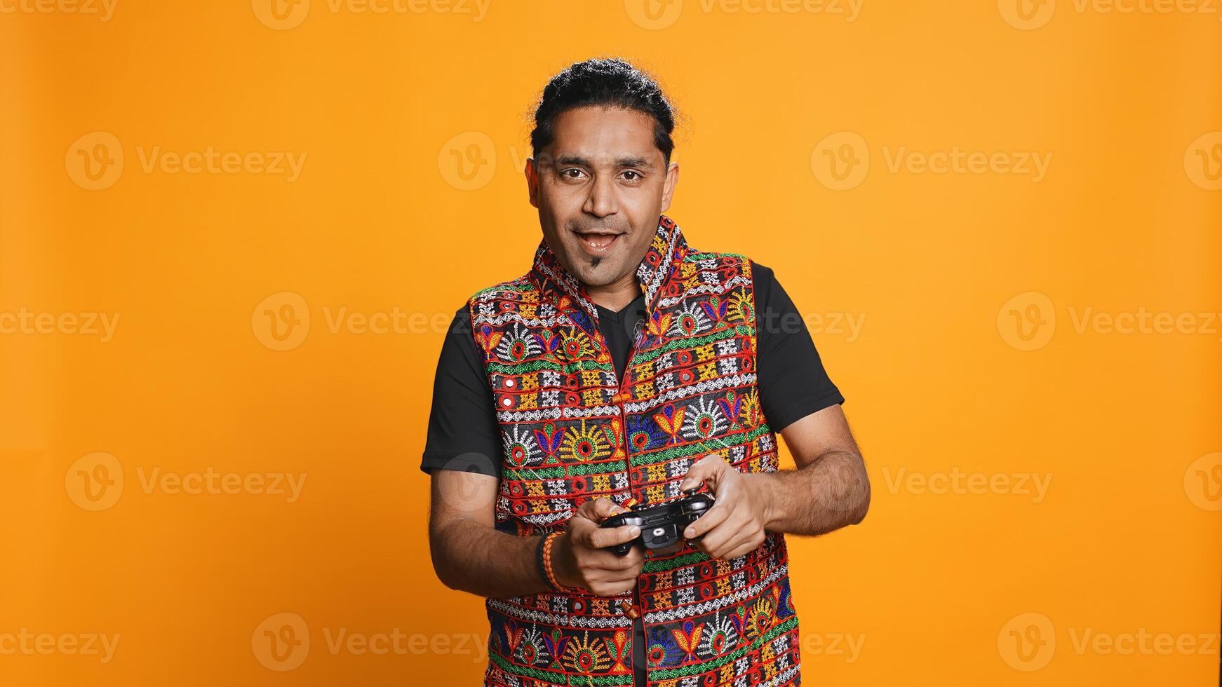 Cheerful indian gamer showing thumbs up sign gesturing, holding controller, playing videogames. Joyful person enjoying games, doing approving hand gesture, studio background, camera B photo