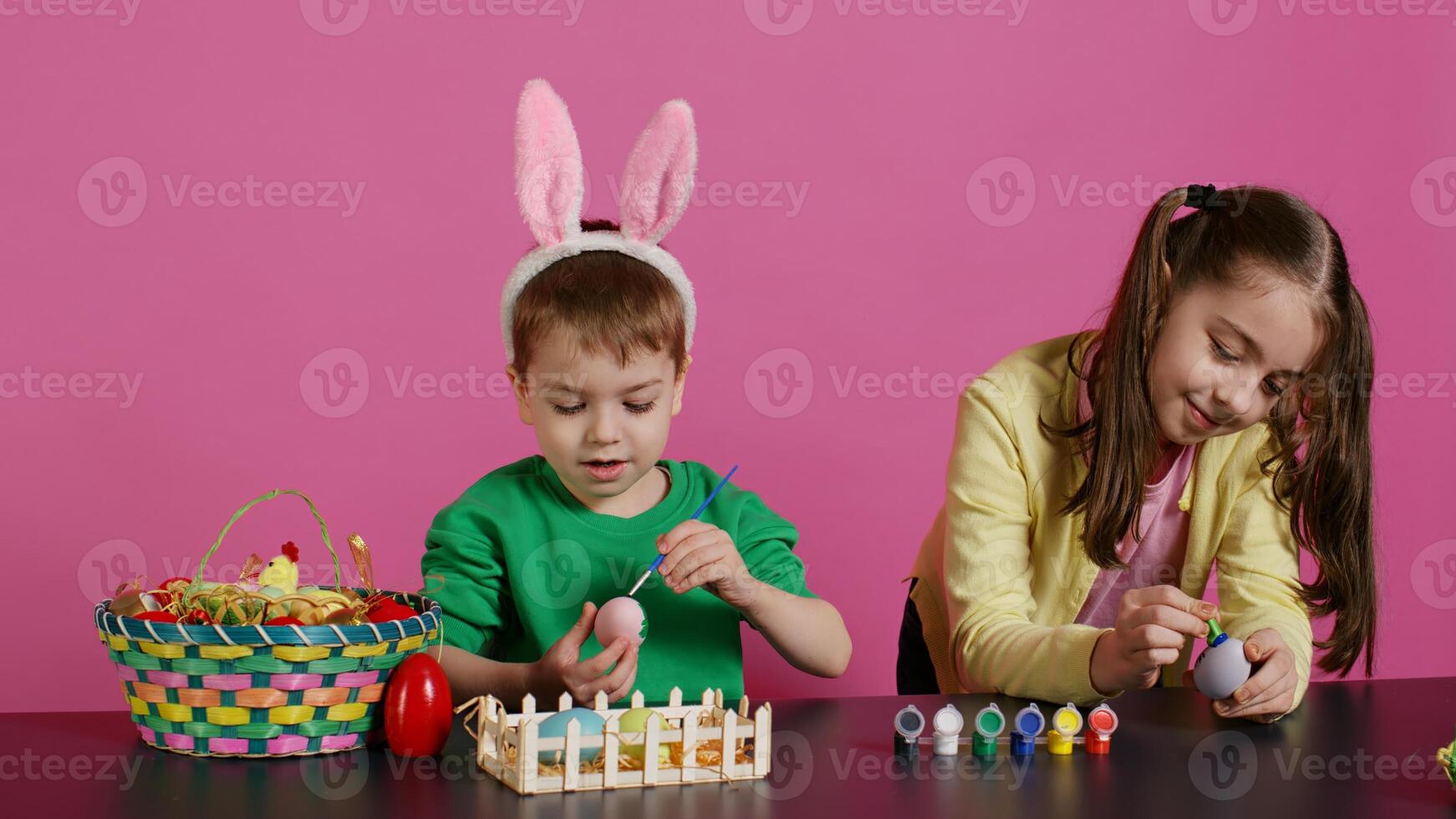 Happy small children using watercolor and stamps to paint eggs in time for easter holiday celebration, pink background. Joyful little siblings decorating ornaments with creative designs. Camera B. photo