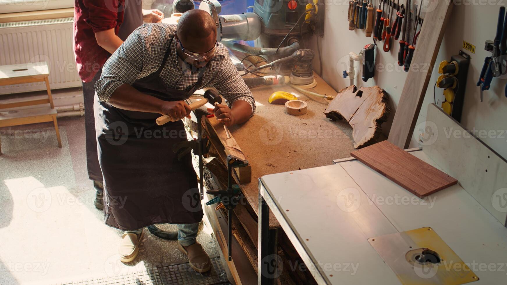 Sculptor shaping raw timber using chisel and hammer in carpentry shop with coworker cleaning workbench in background. BIPOC man making wood sculptures, engraving plank with tools, camera B photo