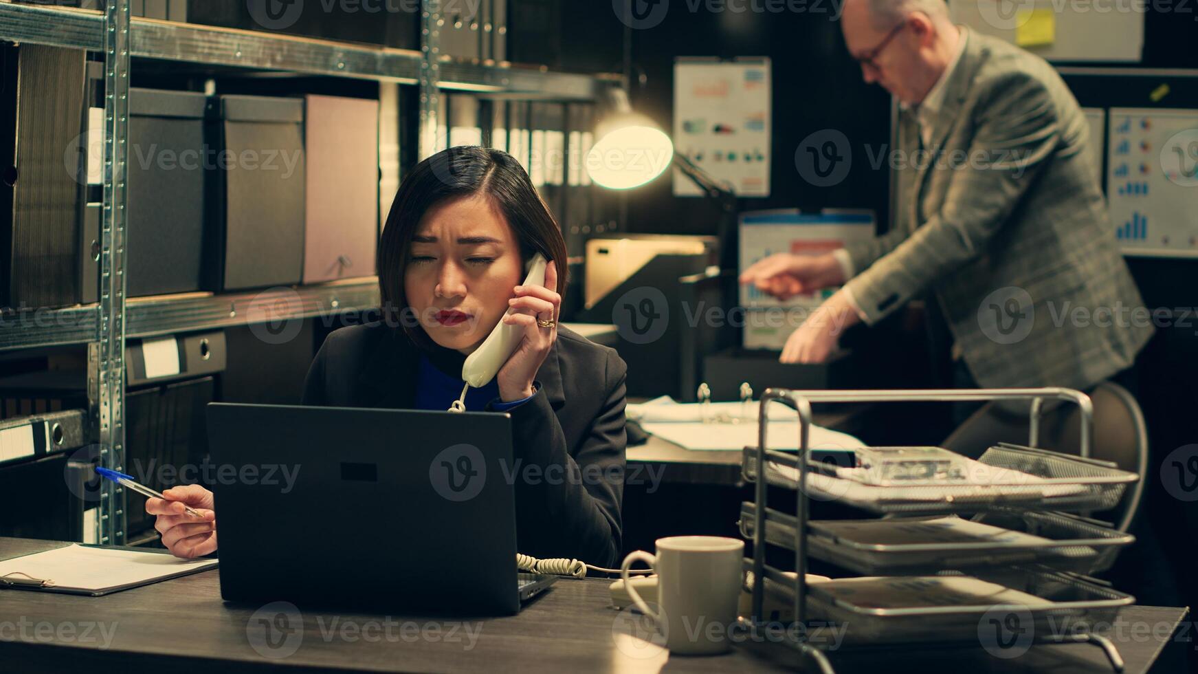 Law officer verifying details for the private investigation, working in archive room. Detective cross checks information on landline phone call, contacting people from records. Camera A. photo