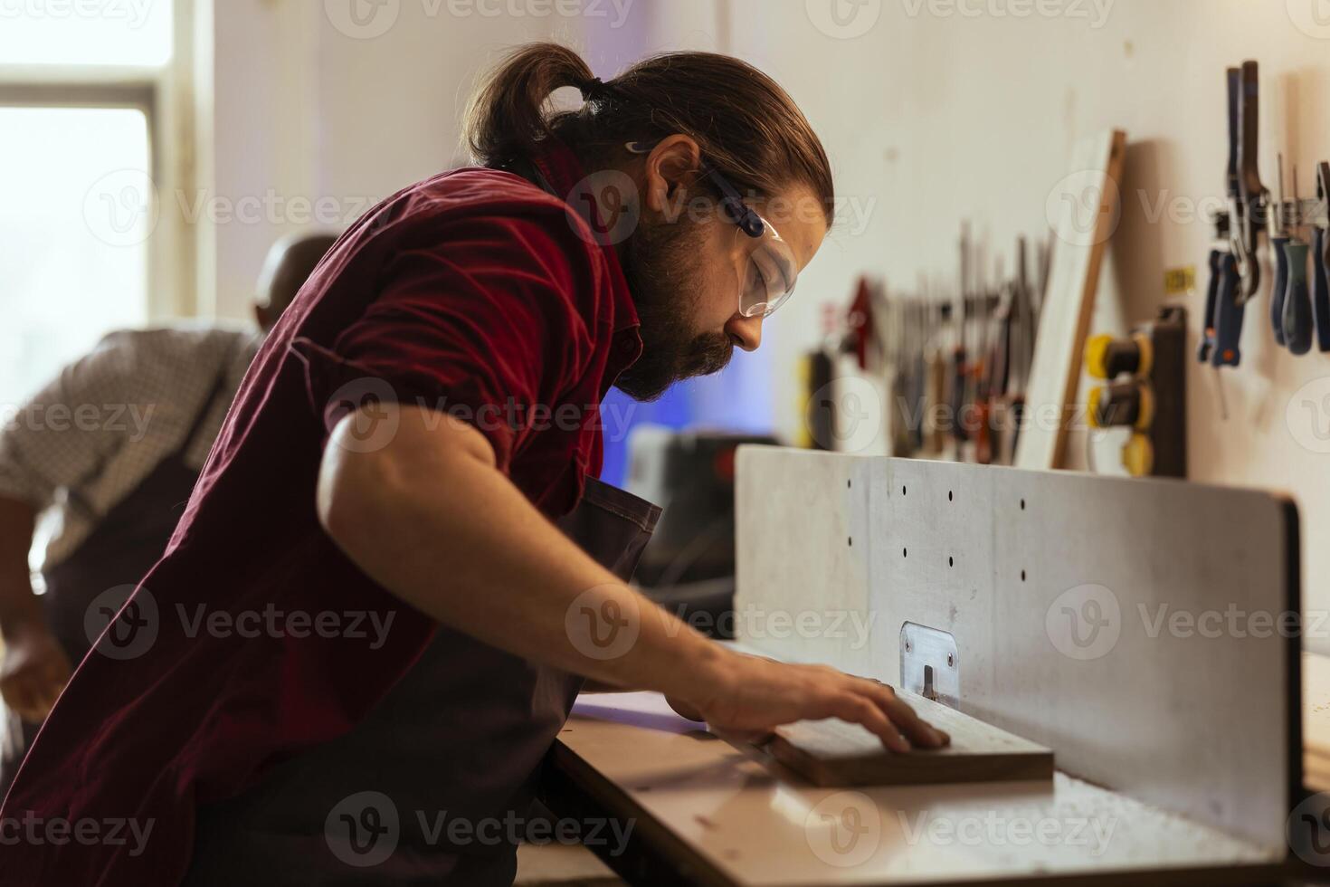 Woodworker operating spindle moulder to create bespoke joinery with coworker in background. Manufacturer and african american apprentice performing tasks on wood shaper, shaping and cutting planks photo