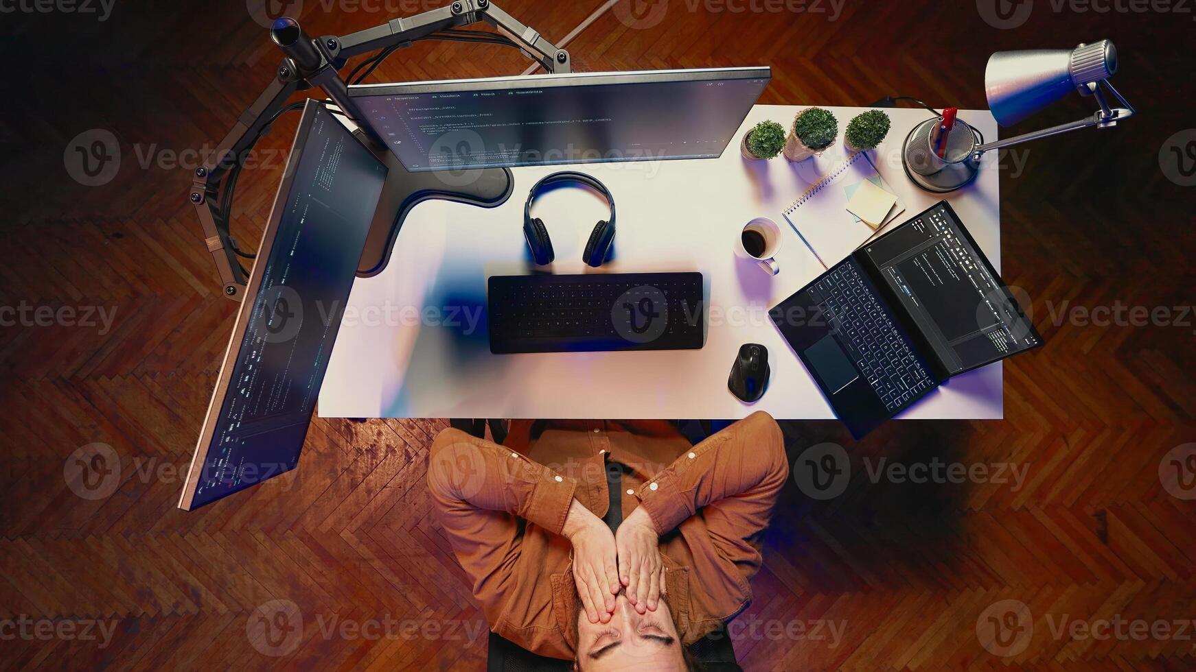 Top down view of fatigued man working overnight at home, rubbing eyes while taking break from coding, feeling sleepy. Tired IT admin exhausted from long screen time, pausing tasks to rest, aerial shot photo