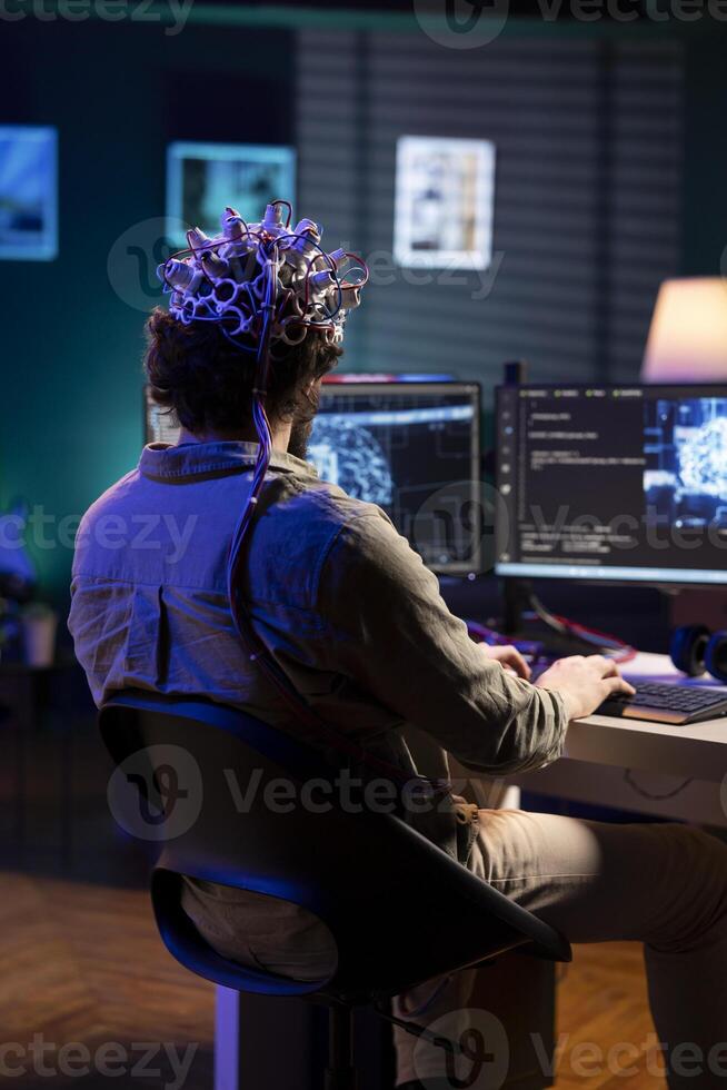 Scientist specializing in artificial intelligence uploading his consciousness into computer, using EEG headset. Man at desk in apartment using high tech device to create link between brain and PC photo