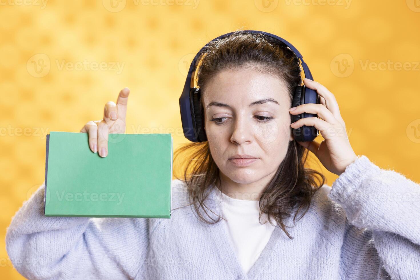 Woman holding book, having fun and listening music, isolated over studio background. Relaxed person with novel in hand enjoying leisure time, hearing songs through stereo headphones photo