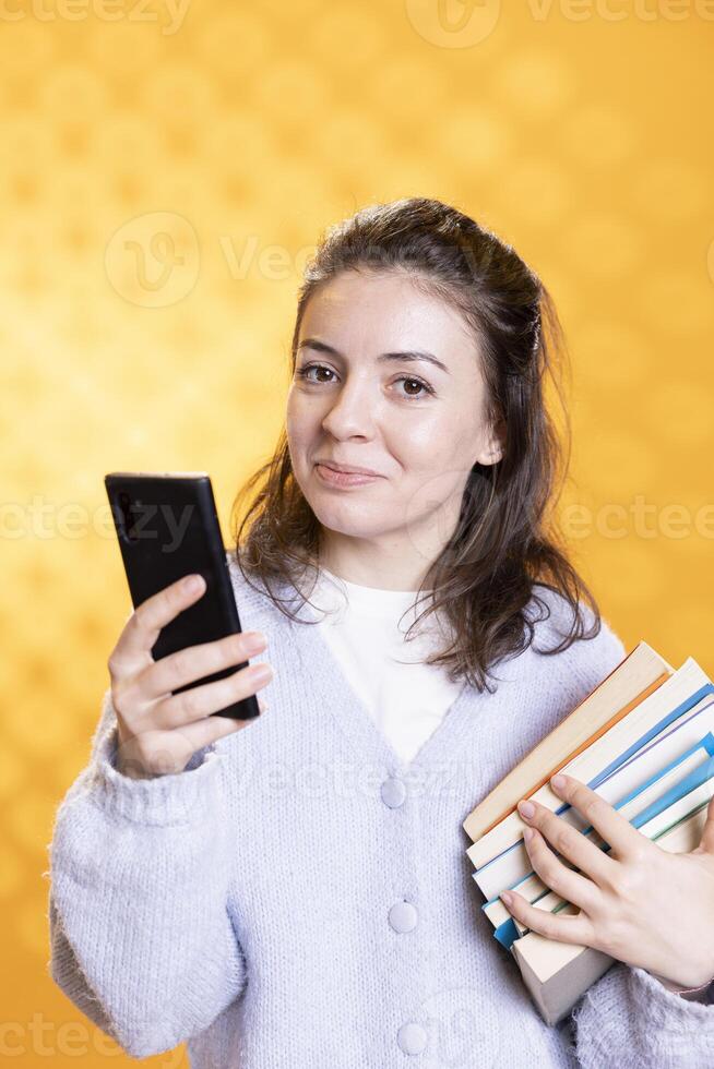 Portrait of smiling woman with stack of textbooks, gathering information for school exam, checking phone screen, studio background. Jolly person holding pile of books, reading messages on smartphone photo