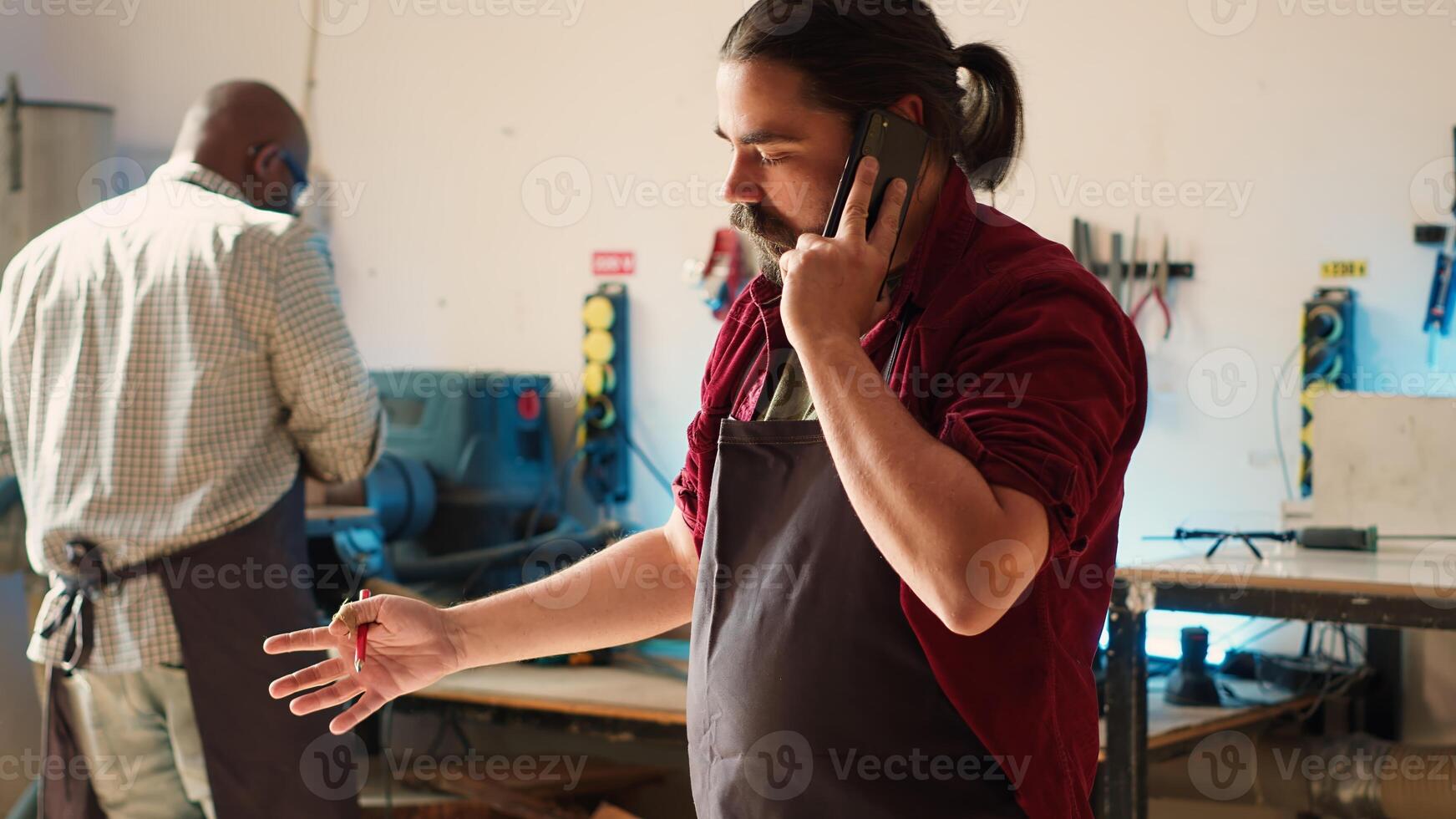 Cabinetmaker receiving telephone call from client commissioning furniture, writing down info. Carpenter in assembly shop drawing schematics, talking on smartphone with customer about order, camera A photo