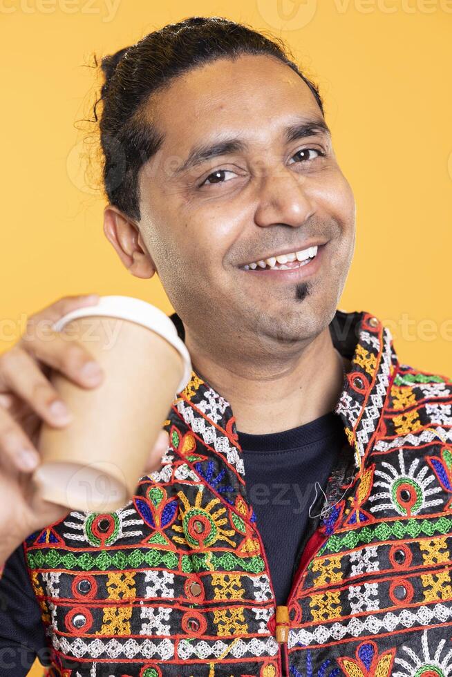 Portrait of ecstatic man wearing traditional indian clothes sipping fresh coffee to wake up and be energized. Merry person with colorful vest drinking hot beverage, studio background, close up shot photo
