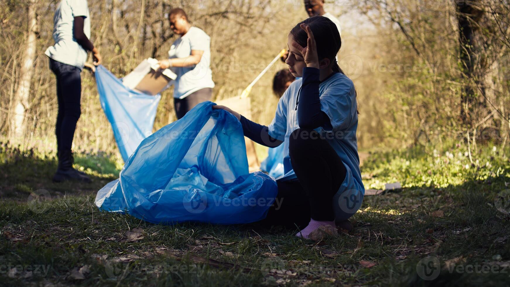 Tired little girl collecting trash and plastic bottles from the forest, helping the volunteers with litter cleanup. Child activist picking up rubbish in a garbage disposal bag. Close up. Camera B. photo