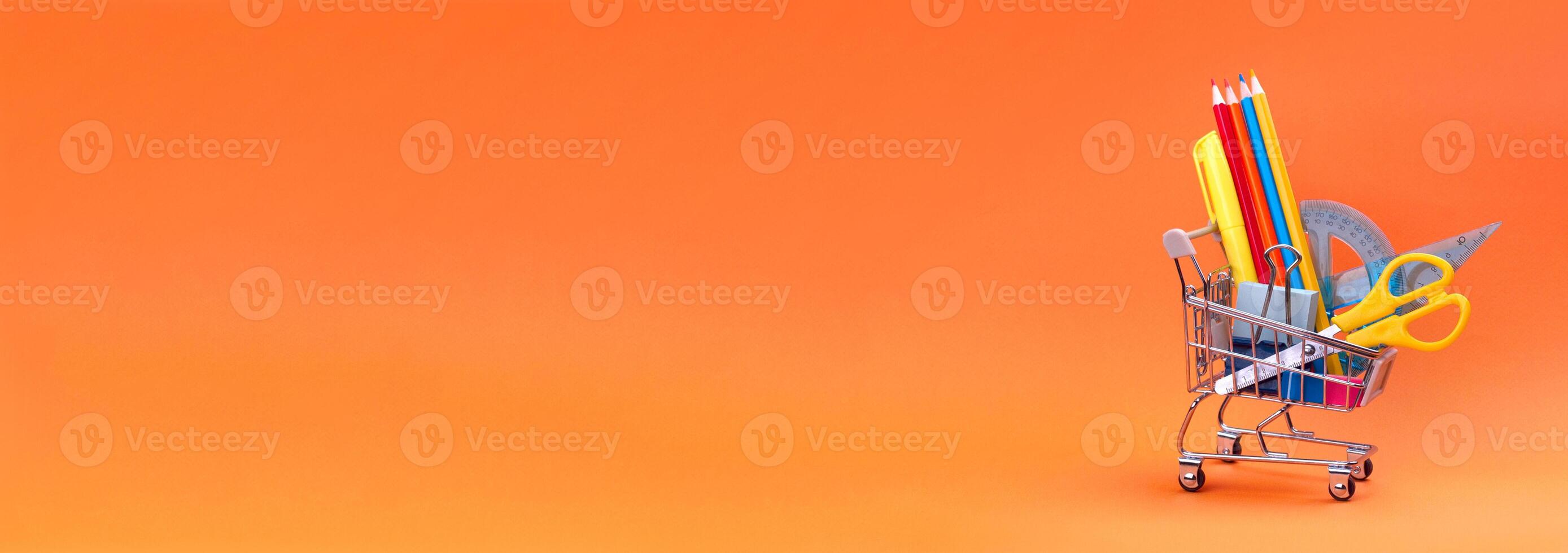 Shopping trolley with stationery items and school supplies on orange background. back to school concept. Preparing for school, buying stationer. banner copy space photo
