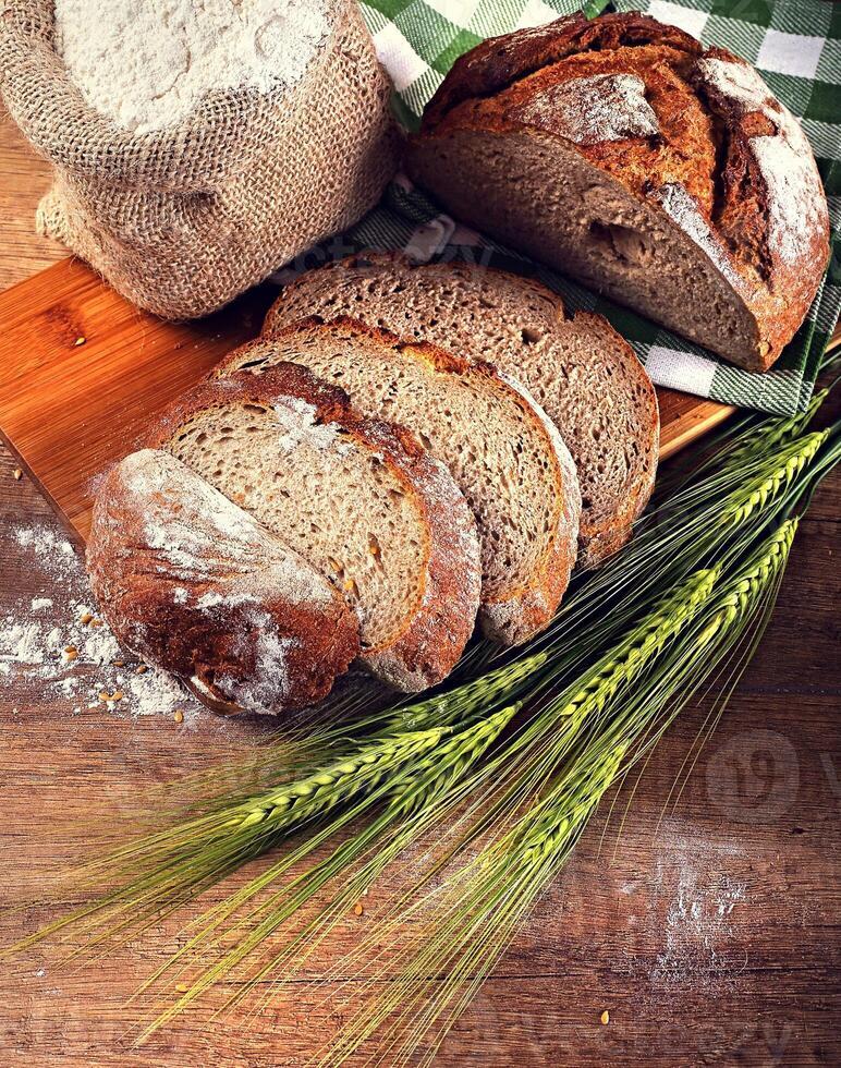 Fresh cereal bread with ears of wheat, with the strewing flour, linseed, bag of flour, bamboo on wooden board, wooden background. photo