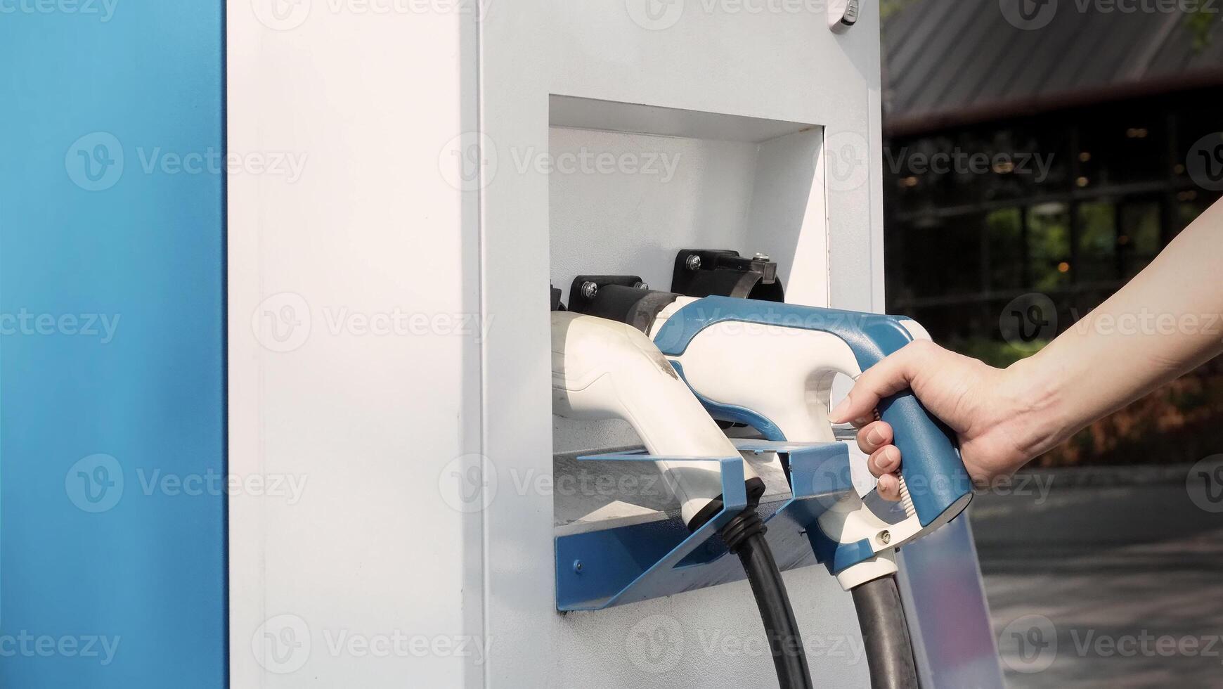 Hand holding EV charging plug in front of camera with blurred background of outdoor. Driver of electric car hold batteries charger plug. Sustainable energy powered electric charging station background photo