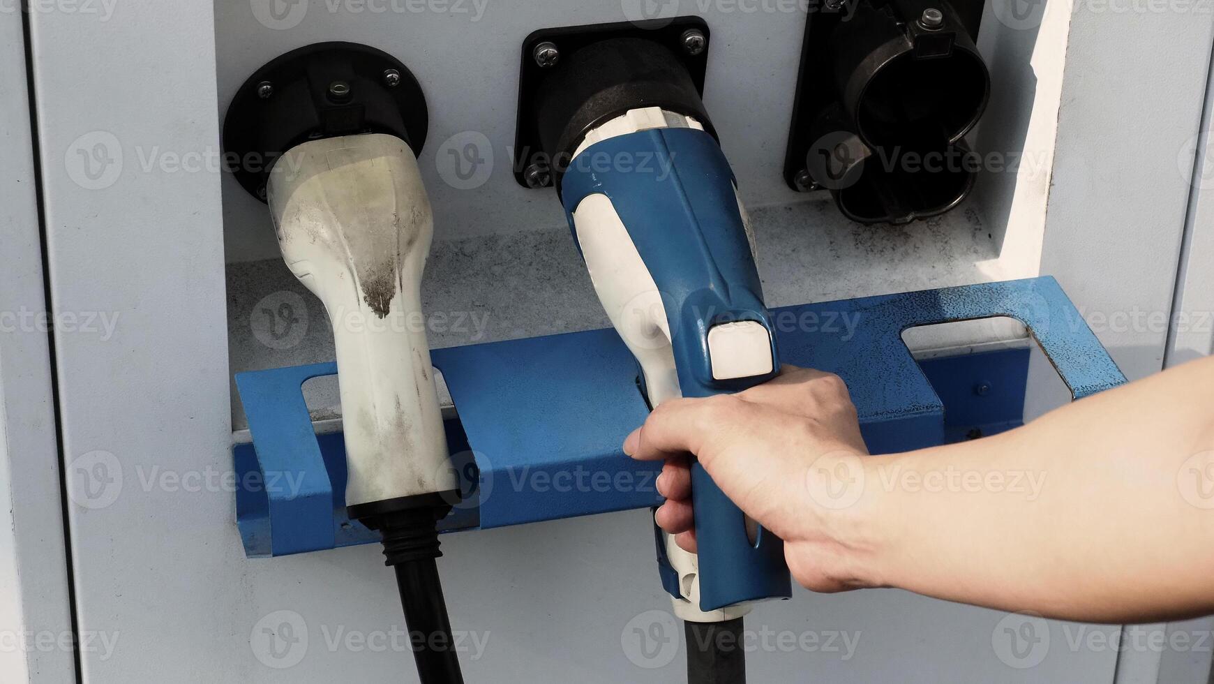 Driver of electric car hold batteries charger plug. Hand holding EV charging plug in front of camera with blurred background of outdoor. Sustainable energy powered electric charging station background photo