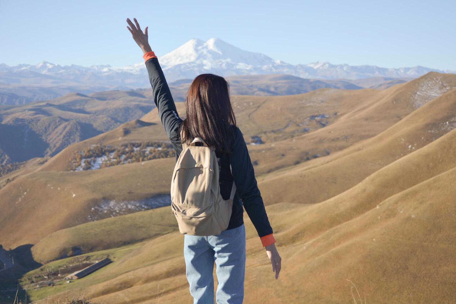 A young brunette woman with a backpack stands against the background of snow-capped Mount Elbrus and looks towards the mountains. photo