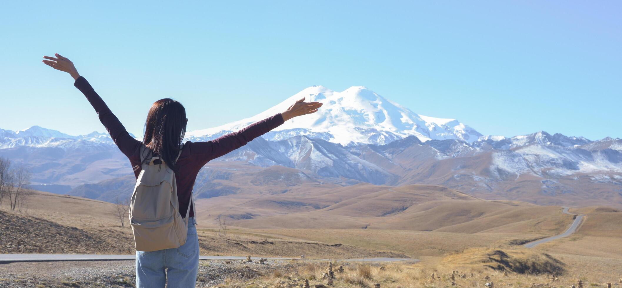 A happy female traveler with a backpack behind her back and her arms raised up admires the snow-covered Elbrus on a clear autumn day. View of Elbrus, North Caucasus, Russia photo